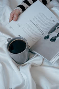 A woman's hand holding a magazine in bed, with a cup of coffee on the sheets, creating a cozy morning vibe.