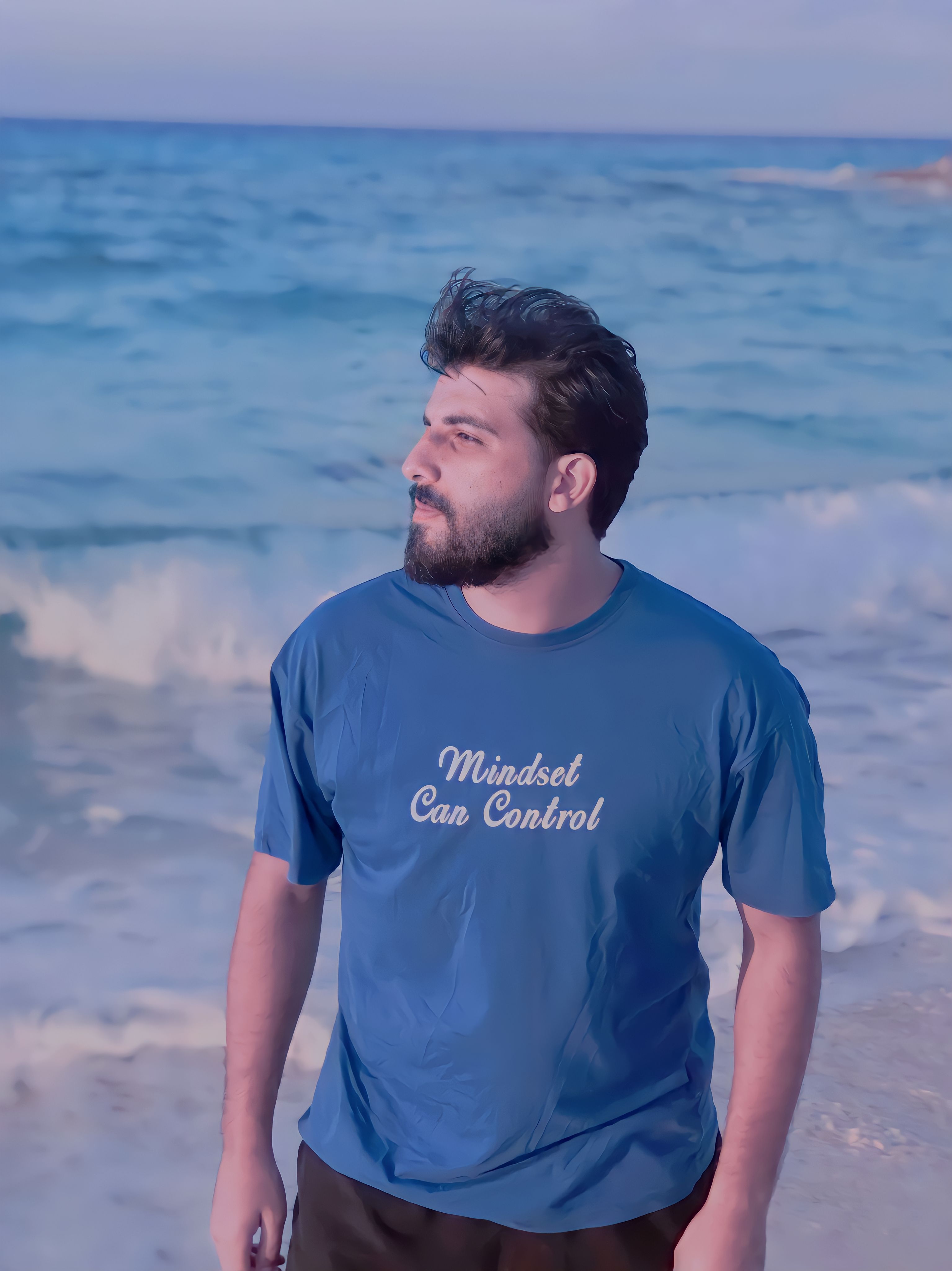 A man in a blue T-shirt with 'Mindset Can Control' text stands by the ocean during the day.