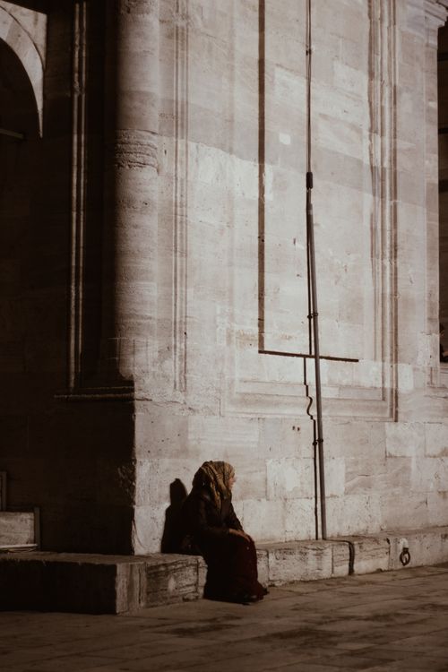 Free A woman wearing a hijab sits quietly against an ancient stone wall at night, casting a long shadow. Stock Photo