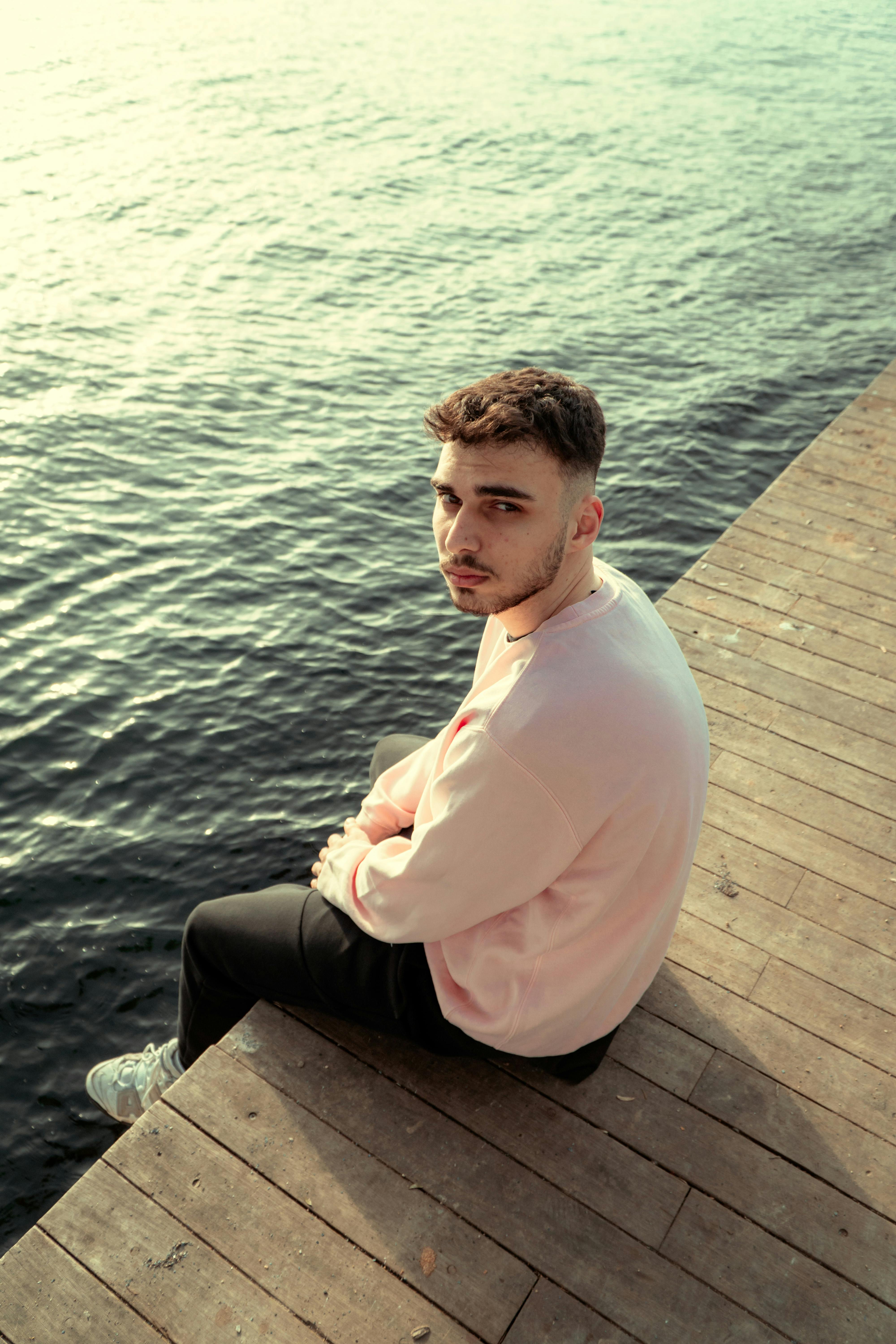 A young man in casual wear sitting on a wooden pier overlooking a calm body of water.