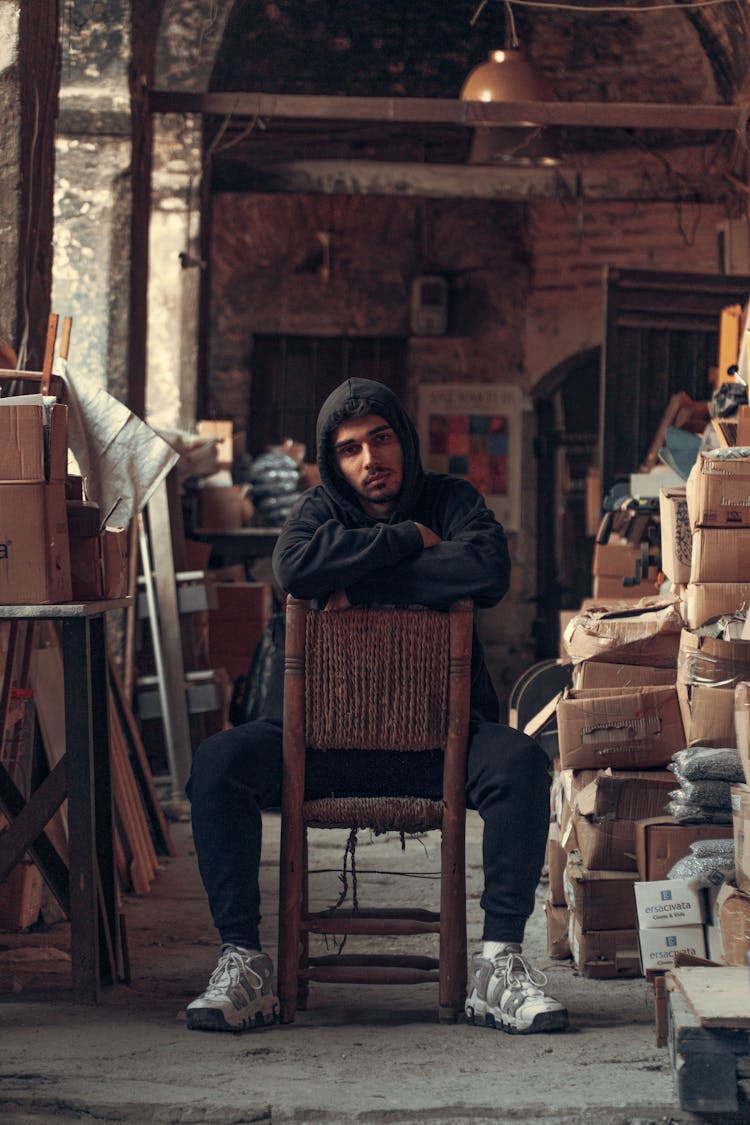 Young Man In A Hoodie Sitting On A Chair Between Stacks Of Boxes 