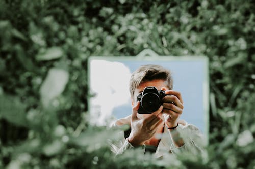 Man Using a Camera Reflecting in a Mirror Standing Between Green Trees