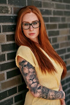 Portrait of a redheaded woman with eyeglasses and tattoos against a brick wall in Frankfurt.