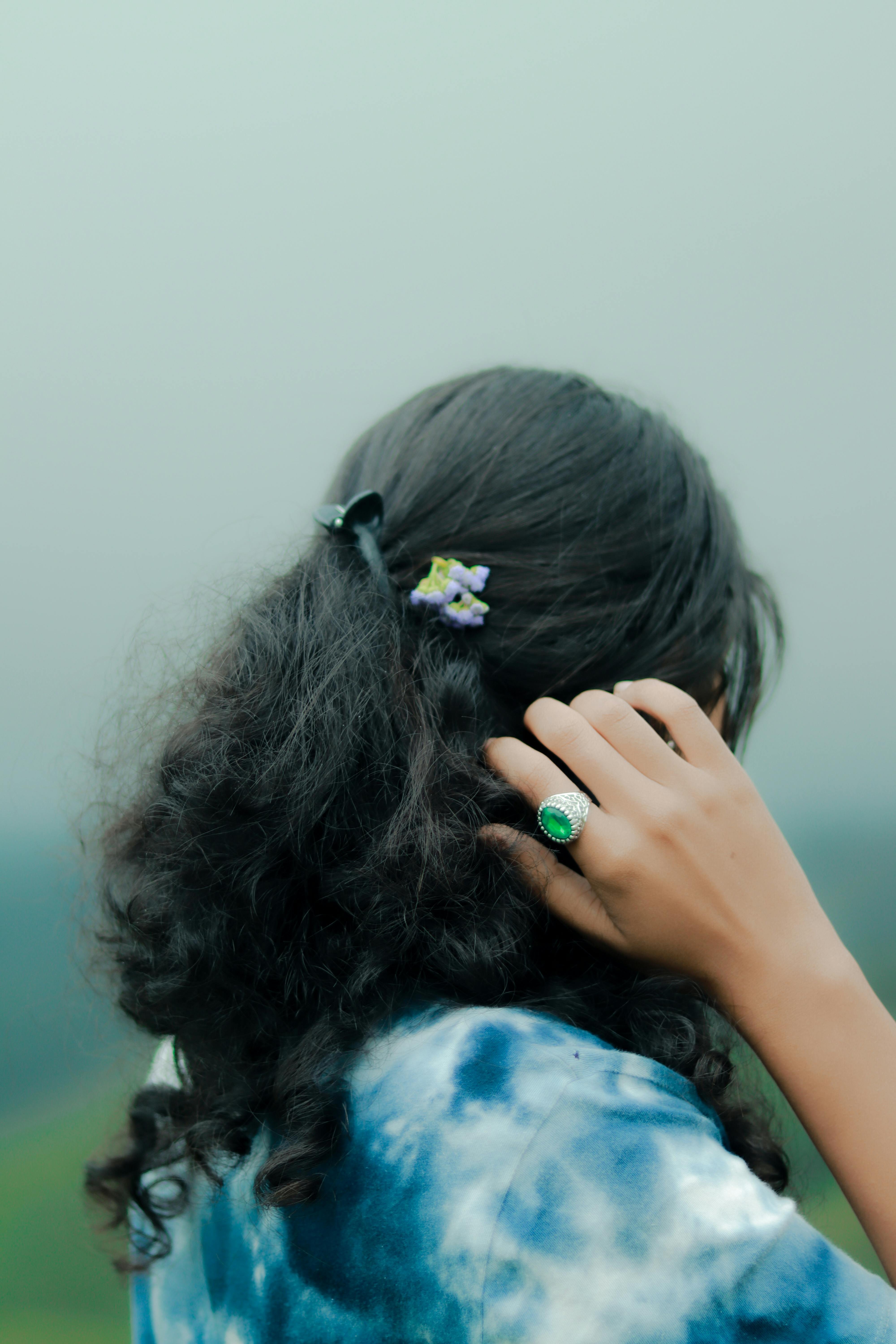 A woman with curly hair adorned with flowers adjusts her hair, showcasing an emerald ring.