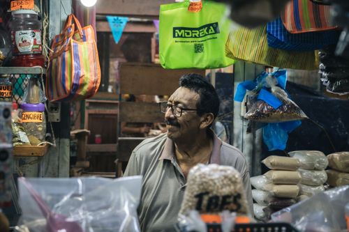 Free Friendly vendor in a local marketplace surrounded by dry goods, bags, and colorful signage. Stock Photo