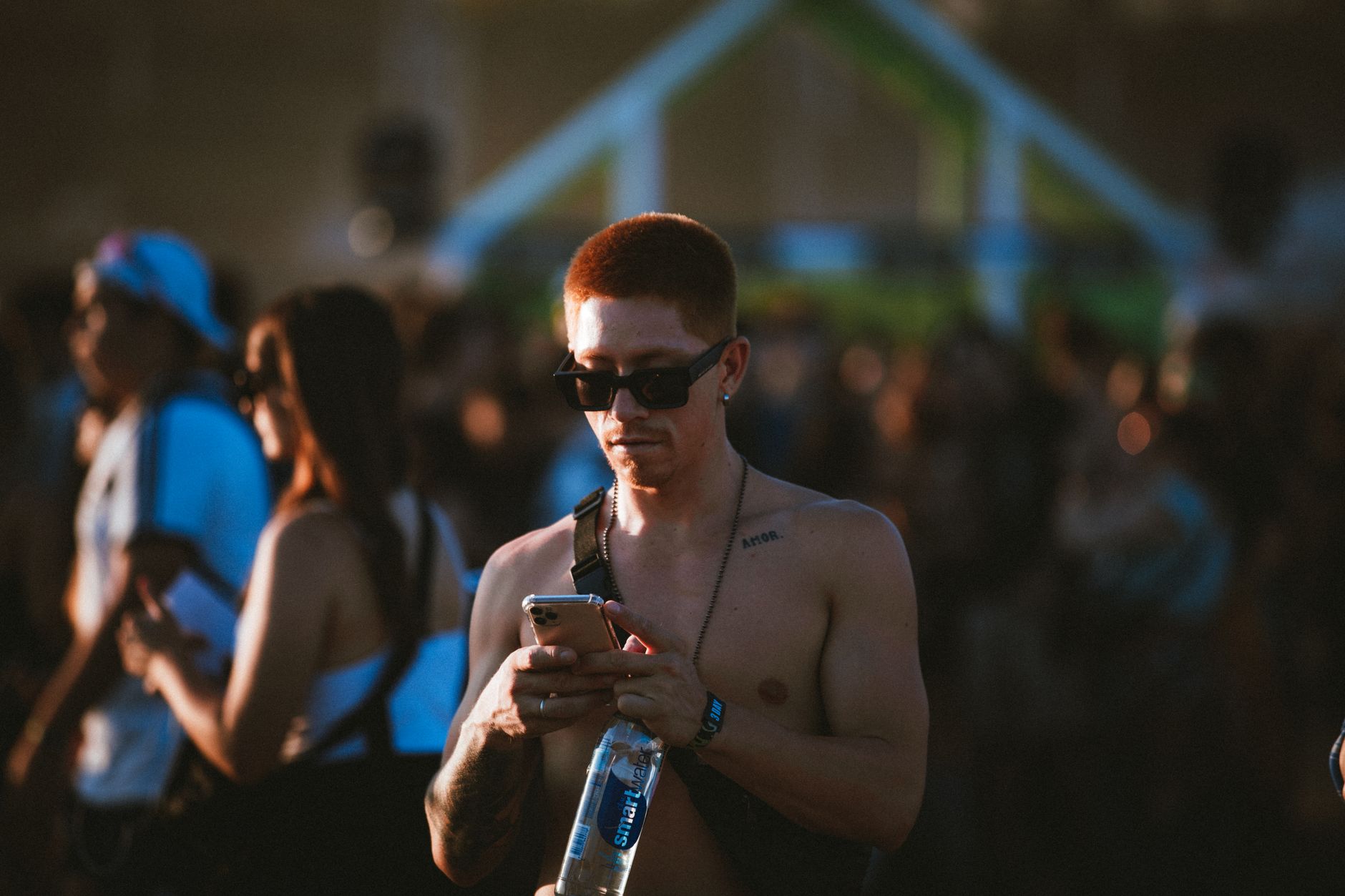 Shirtless man wearing sunglasses at a lively outdoor music festival, checking phone.
