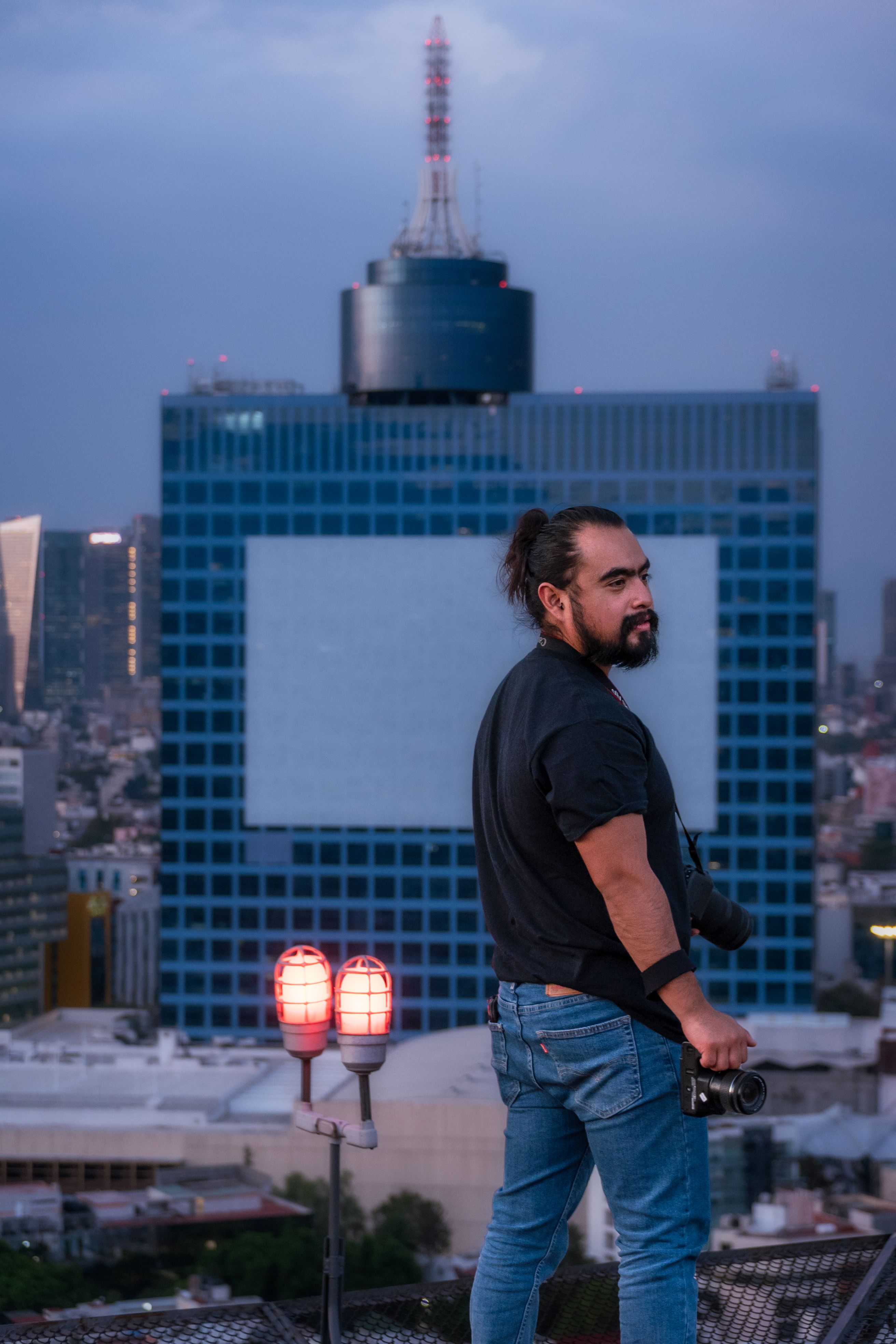 Bearded photographer with camera on a Mexico City rooftop at sunset, against city skyline.