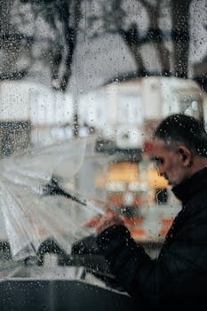 A man holding an umbrella is seen through a rain-soaked window, creating a reflective urban scene.