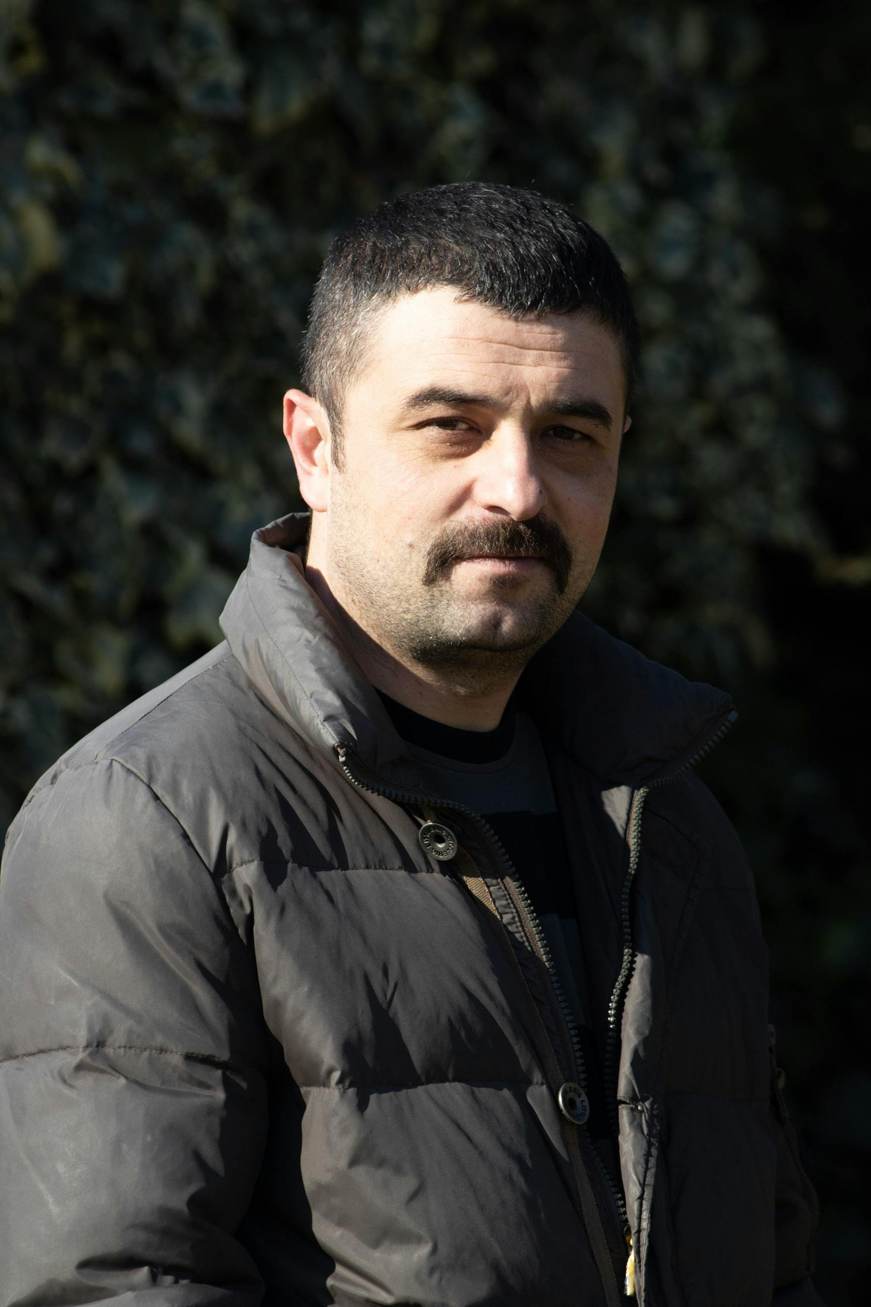 A man with a moustache in a black jacket stands outside in sunlight.
