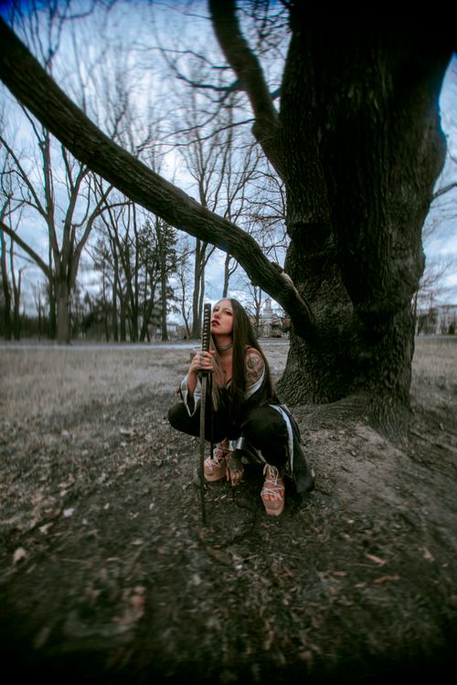 Free A woman in cosplay holding a sword, crouched by a large tree in a park. Stock Photo