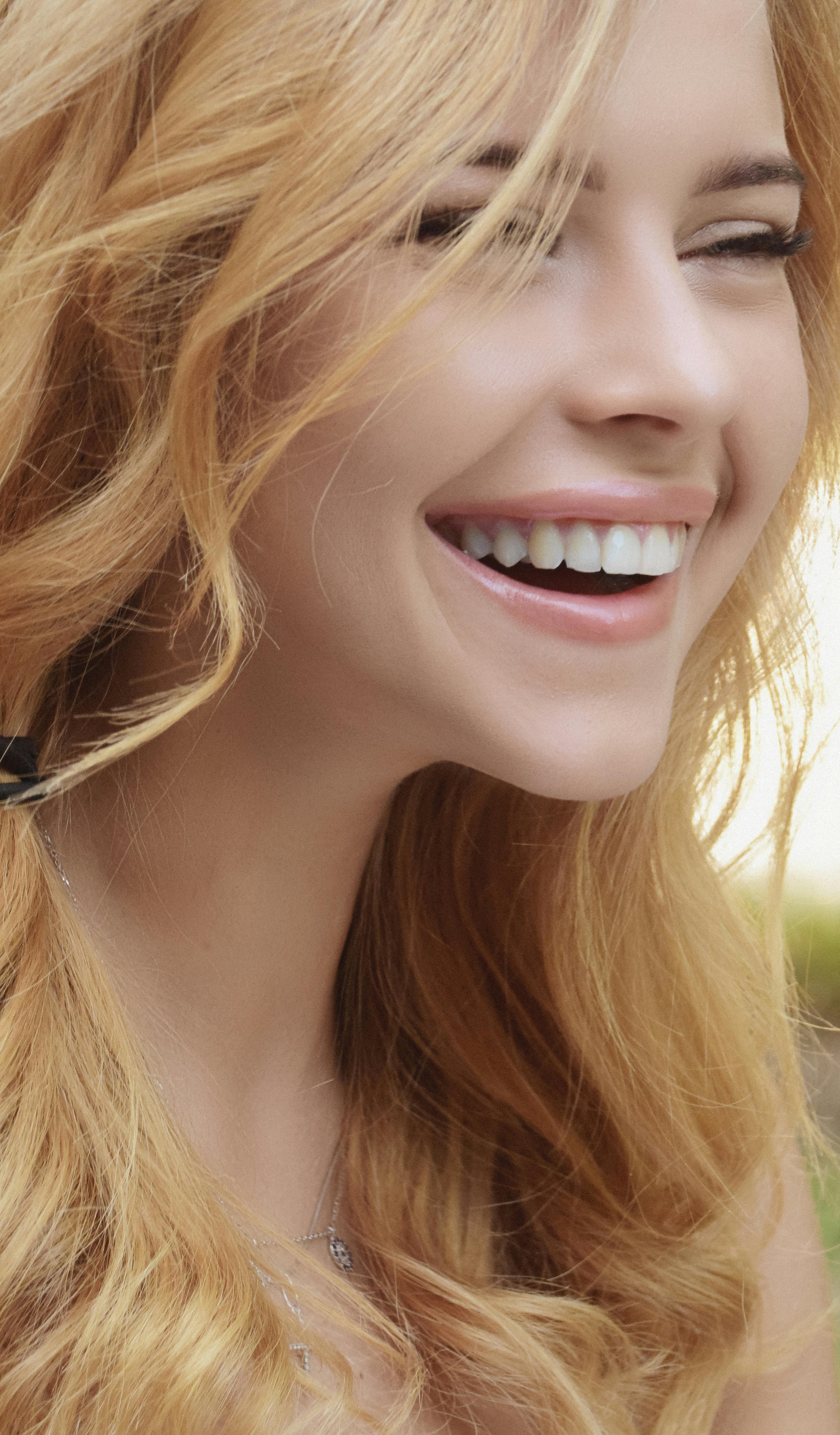 Close-up portrait of a joyful young woman with blonde hair smiling brightly outdoors.