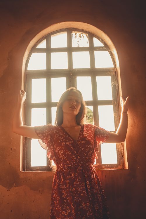 Free A woman in a red floral dress stands peacefully by a sunlit window, creating a warm, serene ambiance. Stock Photo