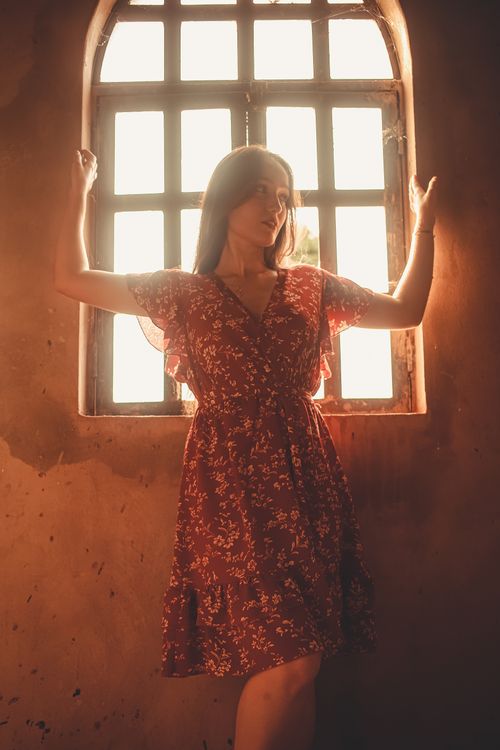 Free A woman in a red floral dress poses gracefully by an arched window with sunlight streaming in. Stock Photo