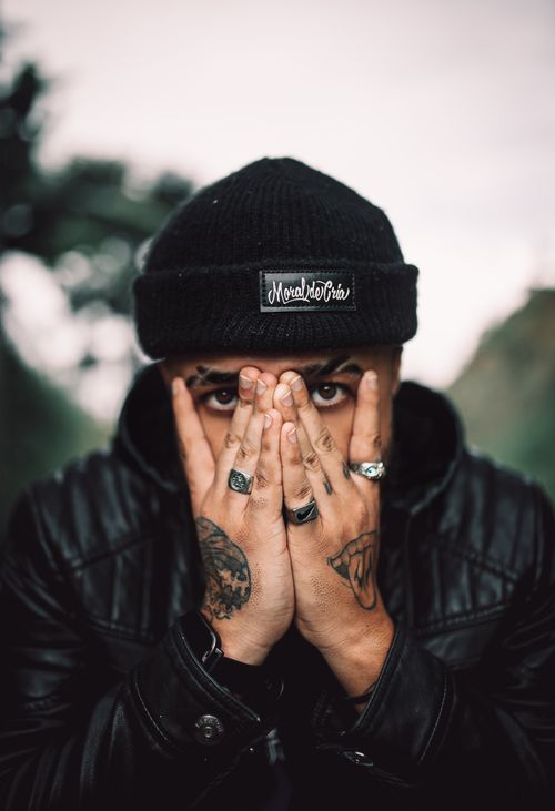 Free Portrait of a man covering face with hands, displaying tattoos and rings in a leather jacket. Stock Photo