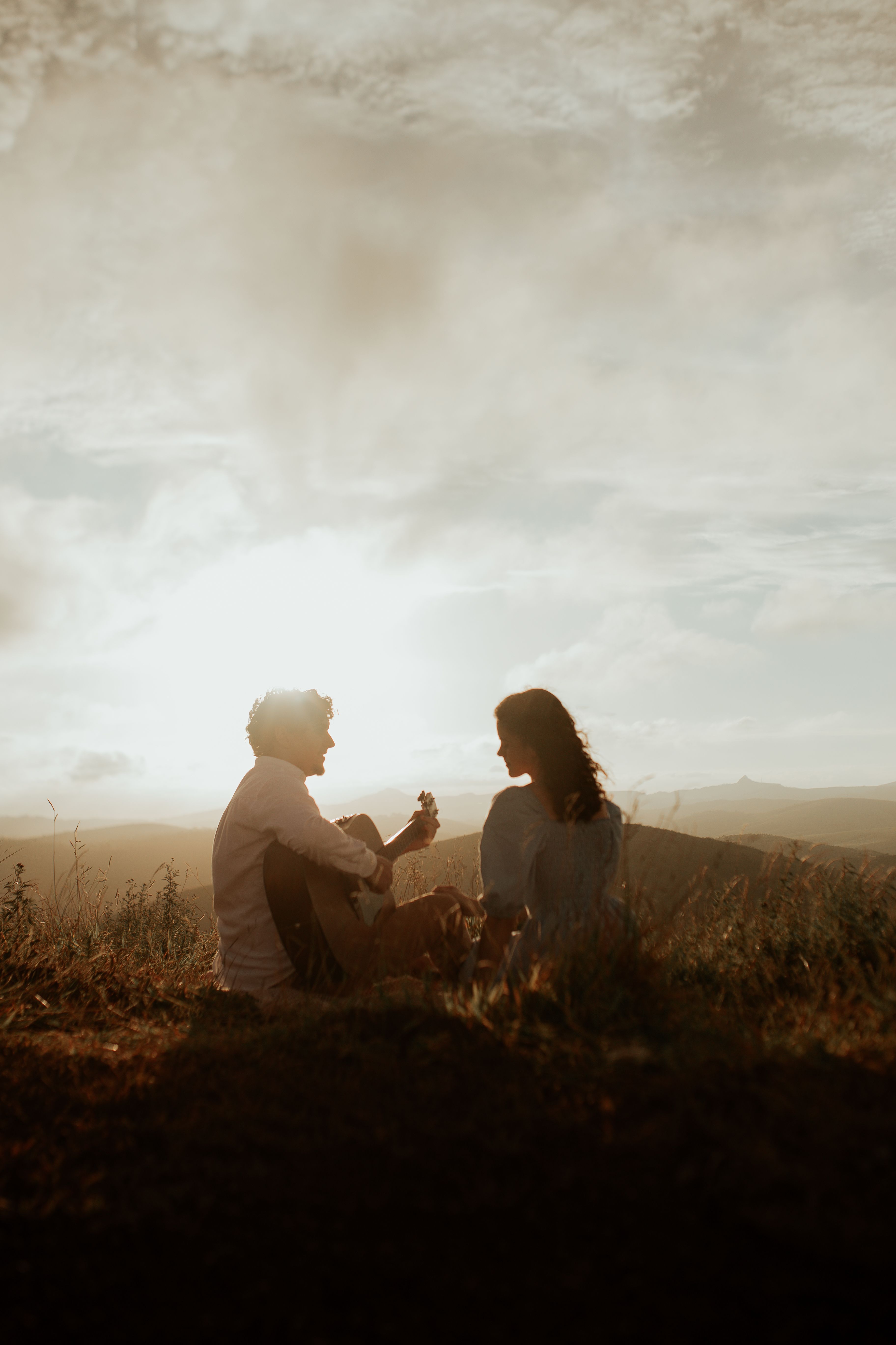 A couple sitting in a field with a guitar, enjoying a romantic sunset.
