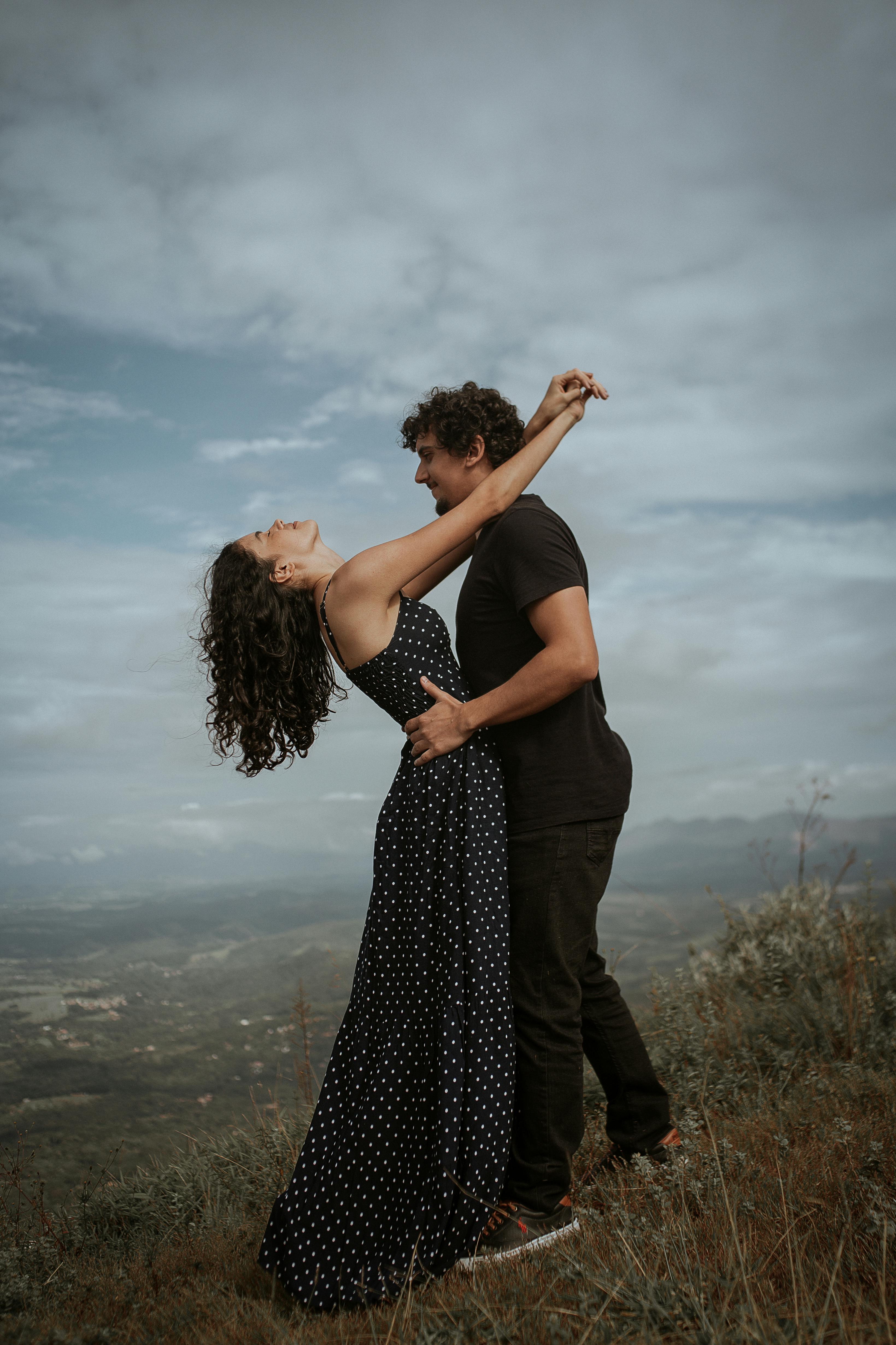 A couple embracing joyfully on a scenic hilltop with a cloudy sky backdrop.