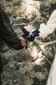 A detailed view of hands holding a floral bouquet against a rustic stone wall.