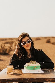 Stylish woman with sunglasses sitting at table with birthday cake in a desert landscape.