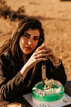 A woman with long dark hair holds a knife near a birthday cake in an outdoor setting.
