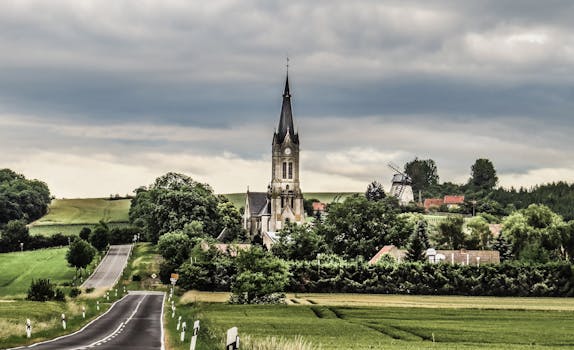 天空多云，郁郁葱葱的田野中矗立着教堂和风车，风景如画