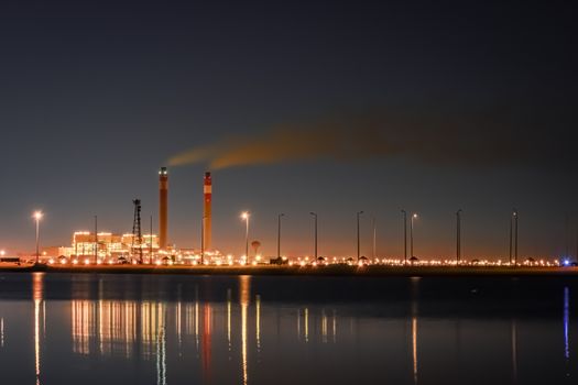 Night view of a lit factory with smoke stacks reflecting on water, symbolizing industrialization.