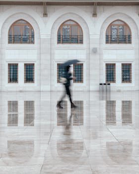 A person with an umbrella walks past a building in İstanbul, Türkiye, reflecting on wet pavement.