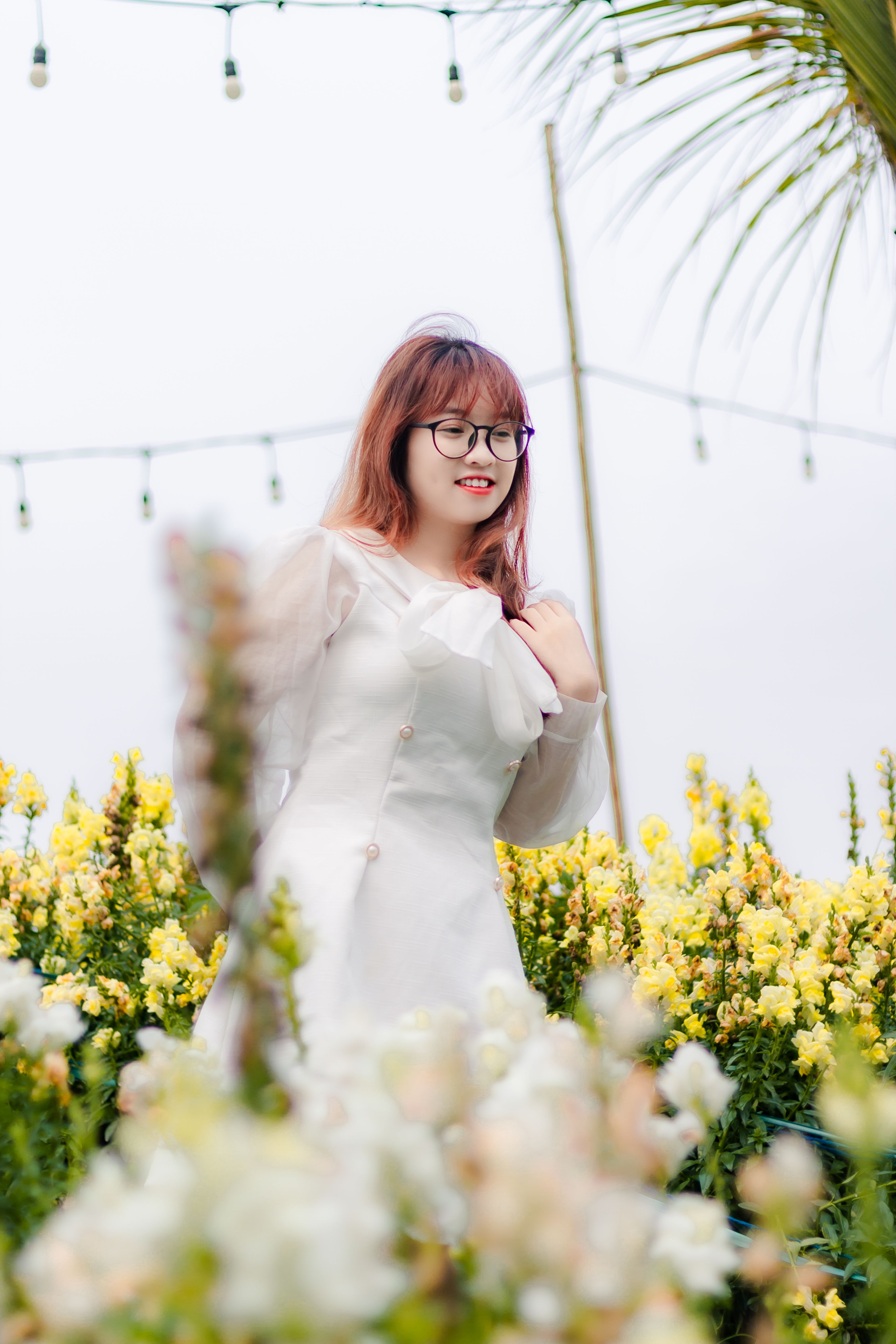 Portrait of a smiling young woman with eyeglasses posing in a lush flower garden, wearing a stylish white dress.