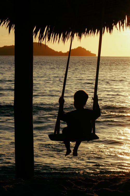 Free Silhouette of a child swinging by the sea during a beautiful sunset scene. Stock Photo