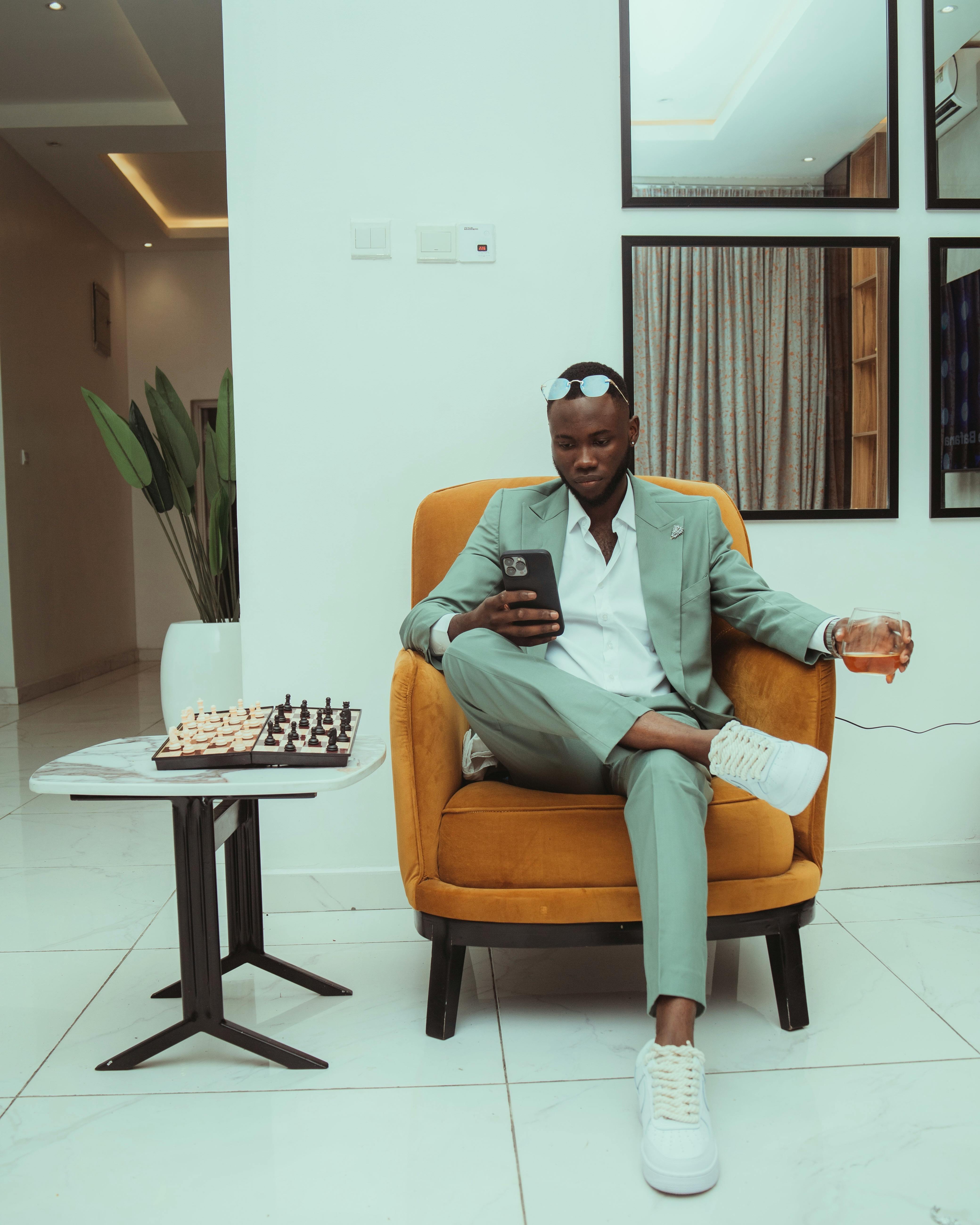 Man in a green suit sitting in an armchair with a cellphone and glass, next to a chessboard.