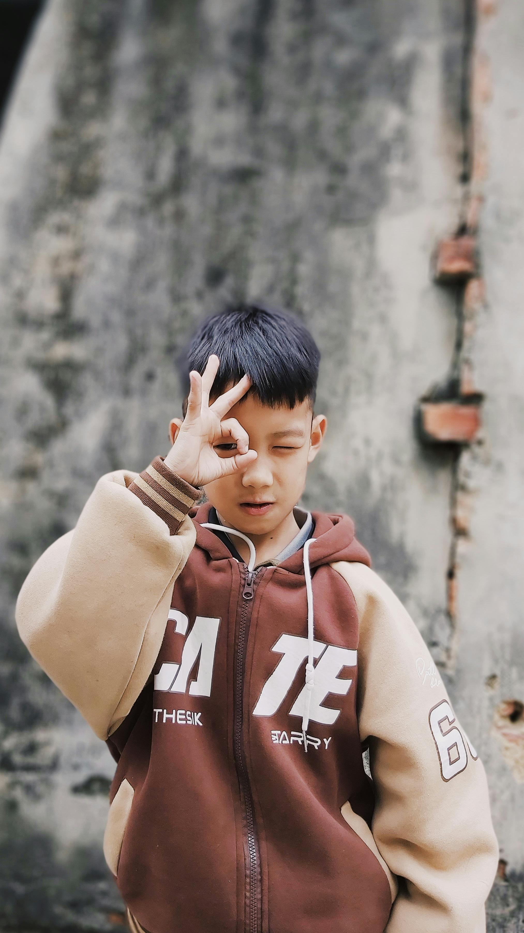 A young boy in a hoodie makes a playful gesture while posing outdoors.