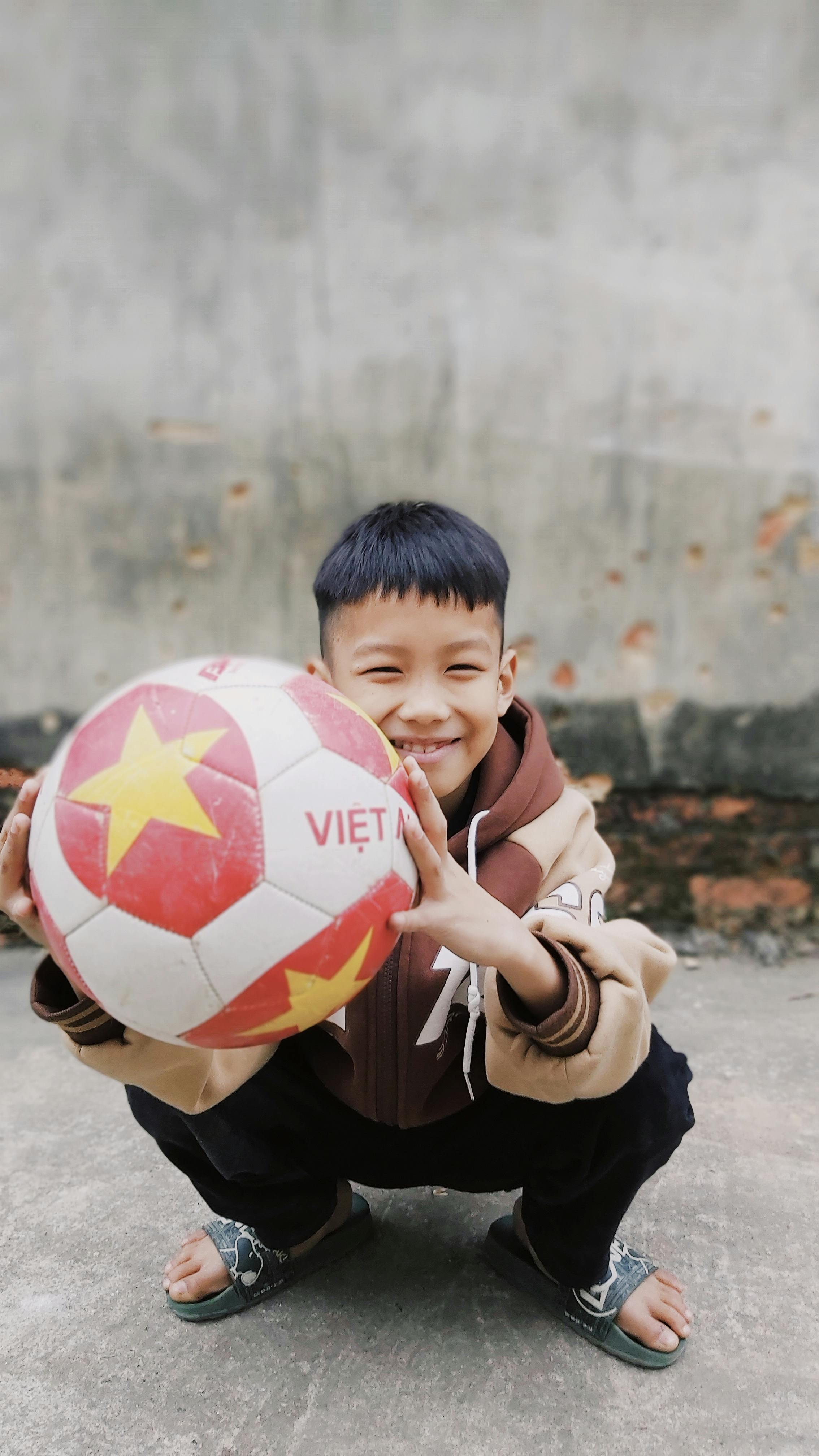 Cheerful Asian boy with black hair crouching outdoors, holding a soccer ball decorated with a Vietnam theme.