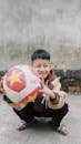 A Little Boy Holding a Ball and Crouching Outside