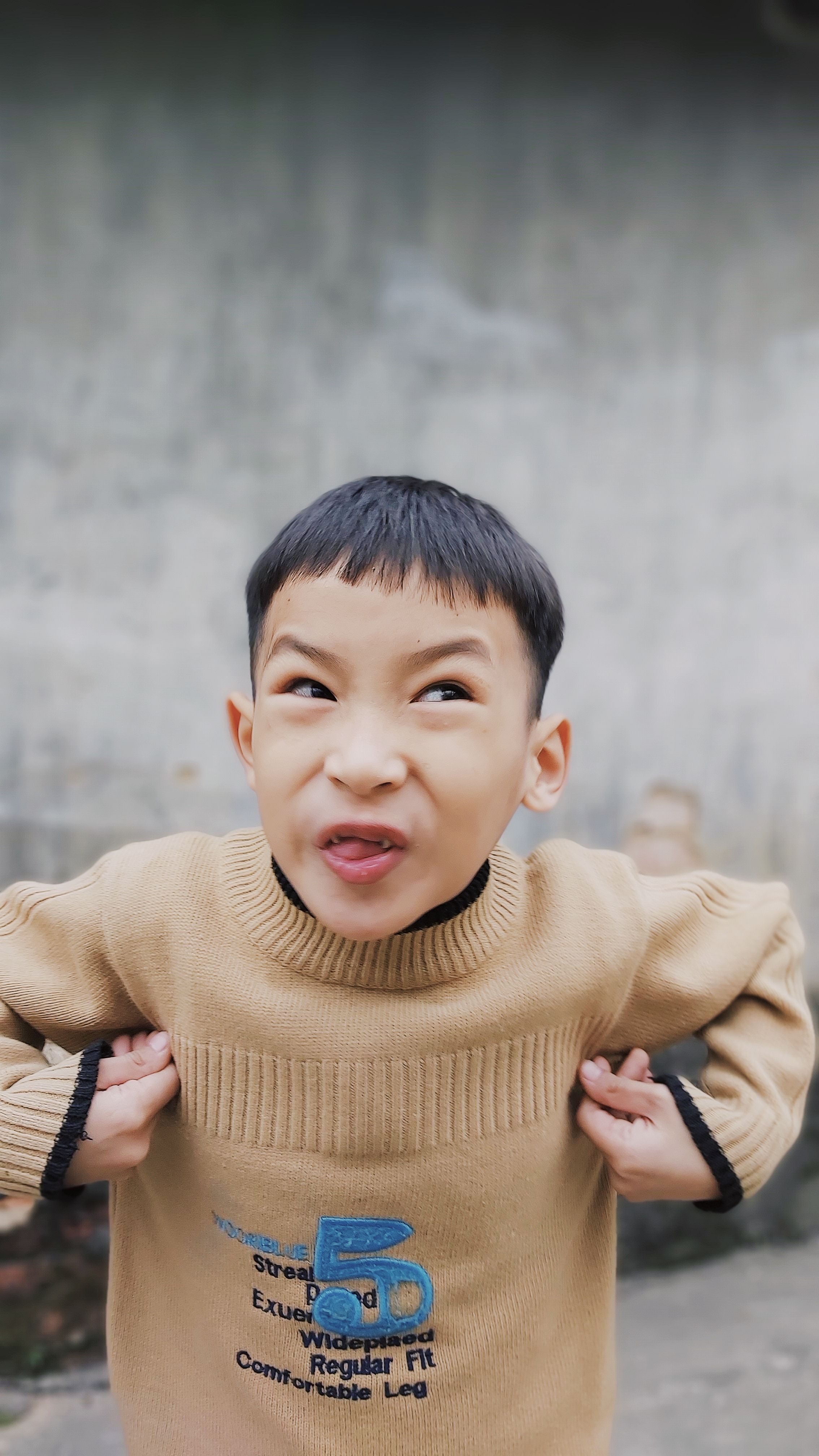 A playful child with black hair makes a funny face while wearing a sweater outdoors.