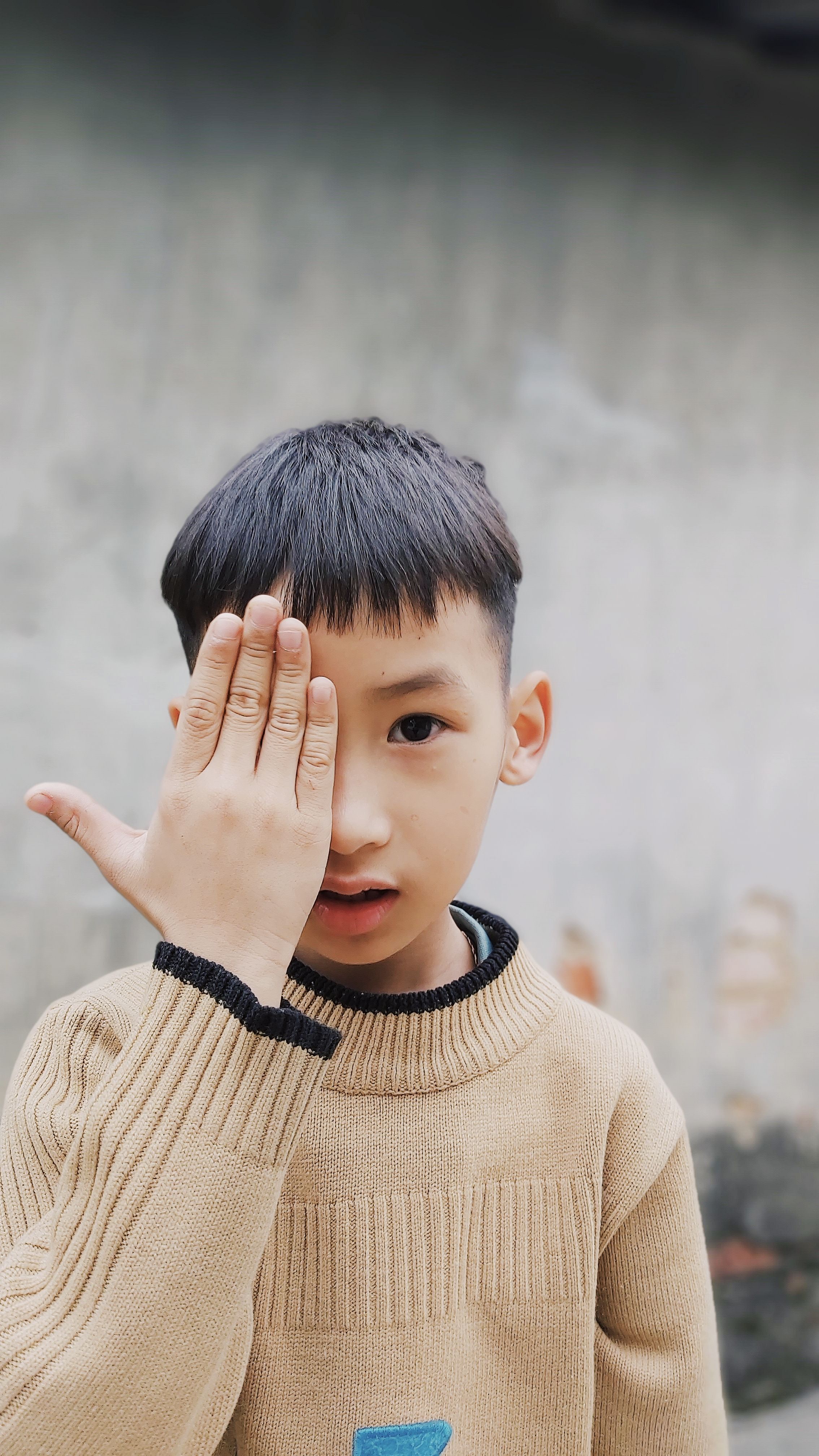 Cute boy with black hair in a brown sweater covering one eye with his hand outdoors.