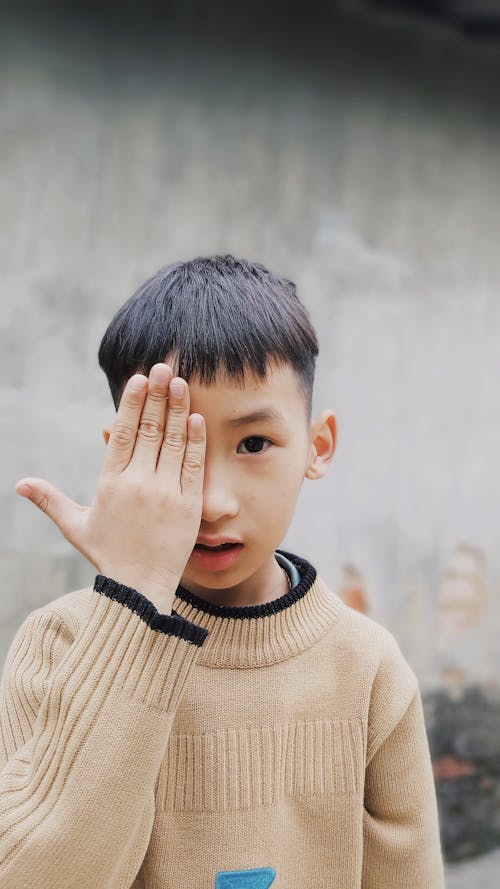 A Little Boy in a Brown Sweater Standing Outside and Covering One of His Eyes with His Hand 