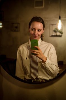 A woman in a white shirt and tie takes a selfie with her cellphone in a warmly lit setting.