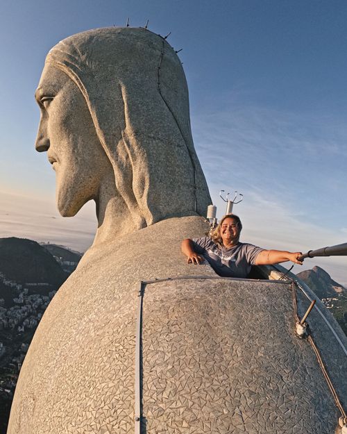 Free Unique close-up view of Christ the Redeemer with a smiling tourist on top of the statue under a blue sky. Stock Photo