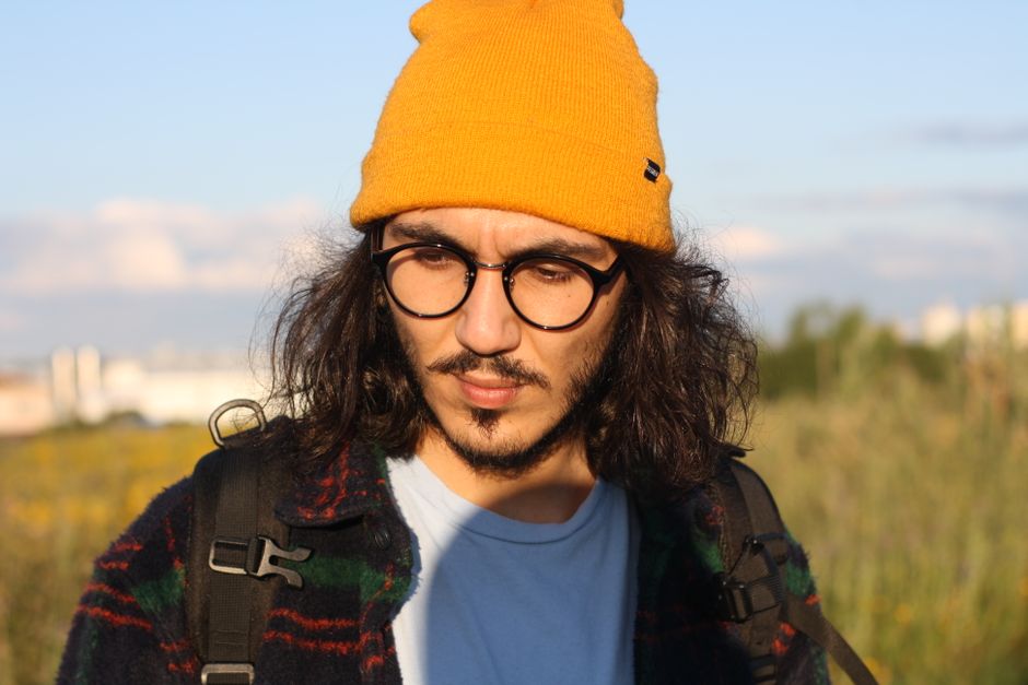 A thoughtful young man with glasses standing outdoors in a sunny field wearing a yellow beanie.
