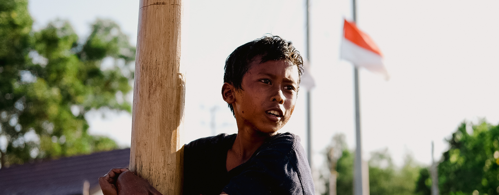A child climbing a wooden pole in an outdoor setting with the Indonesian flag visible in the background.