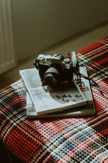 A vintage camera sits on magazines on a patterned blanket in warm sunlight.
