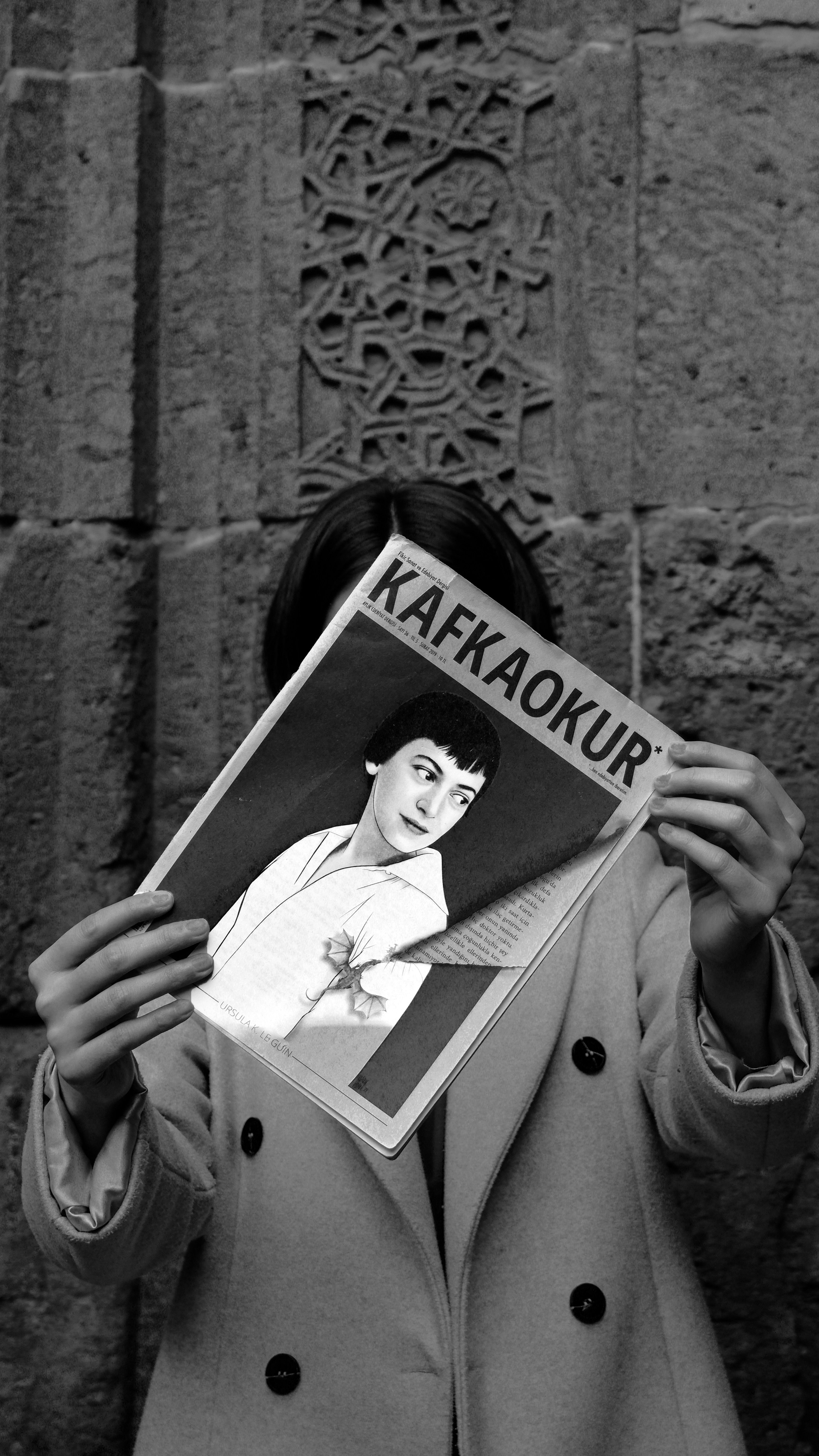 A person holding a Kafkaokur magazine in front of a stone wall, creating a mysterious vibe.