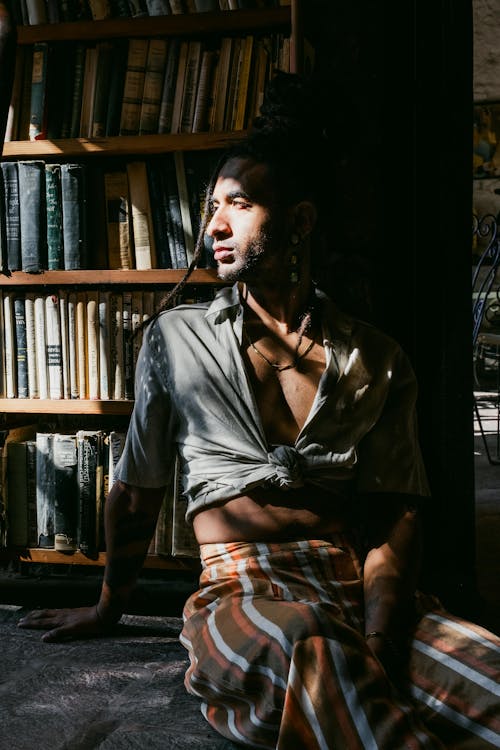 Young Man with Dreadlocks Sitting on the Floor and Leaning against a Bookcase 