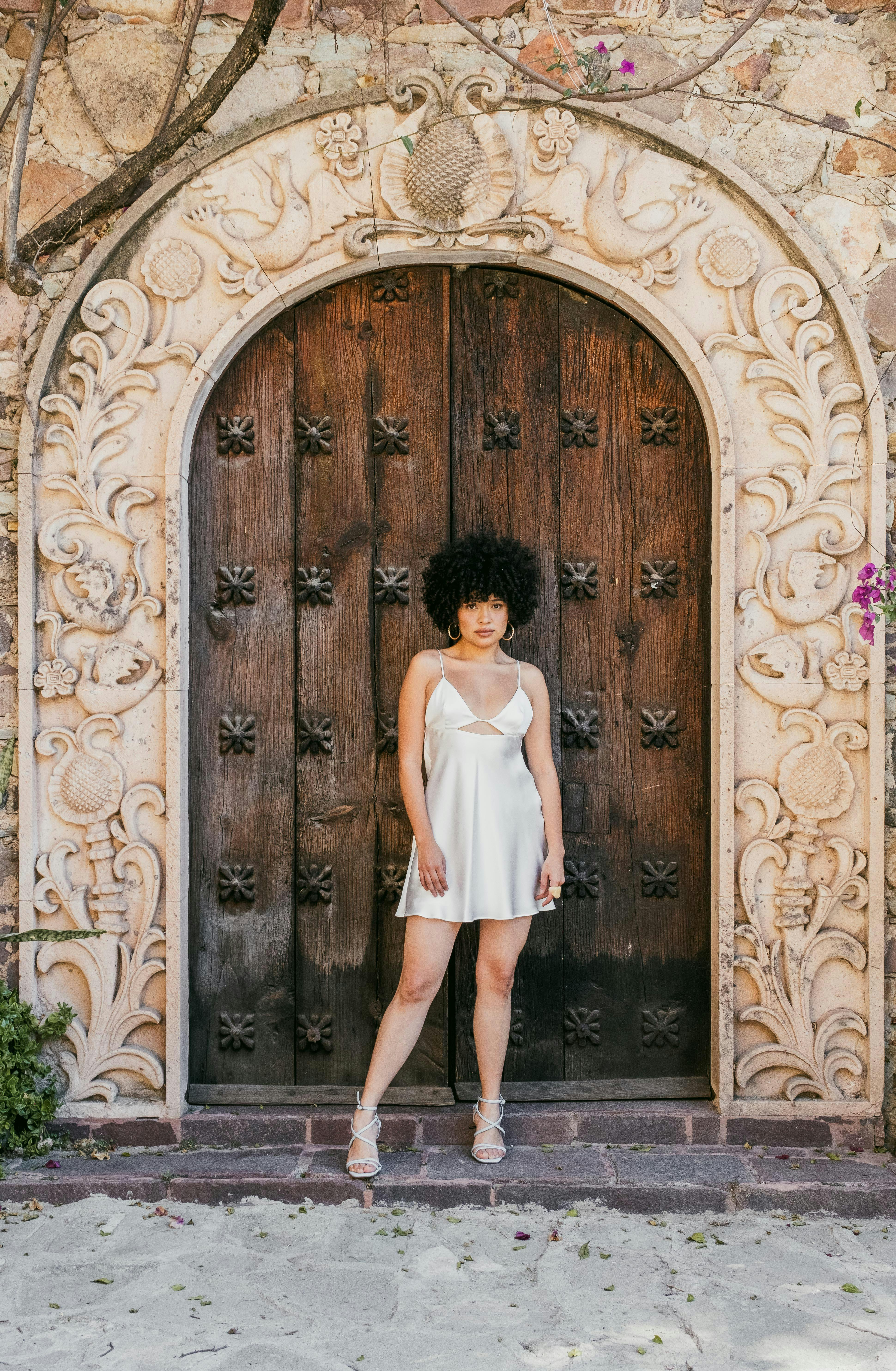 A woman in a white dress poses confidently at an ornate wooden door.