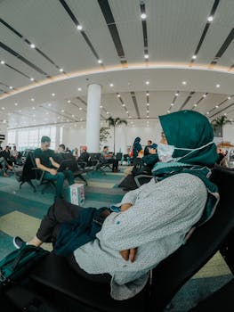 A woman in a hijab and mask patiently waits in a busy airport lounge, showcasing travel and safety.