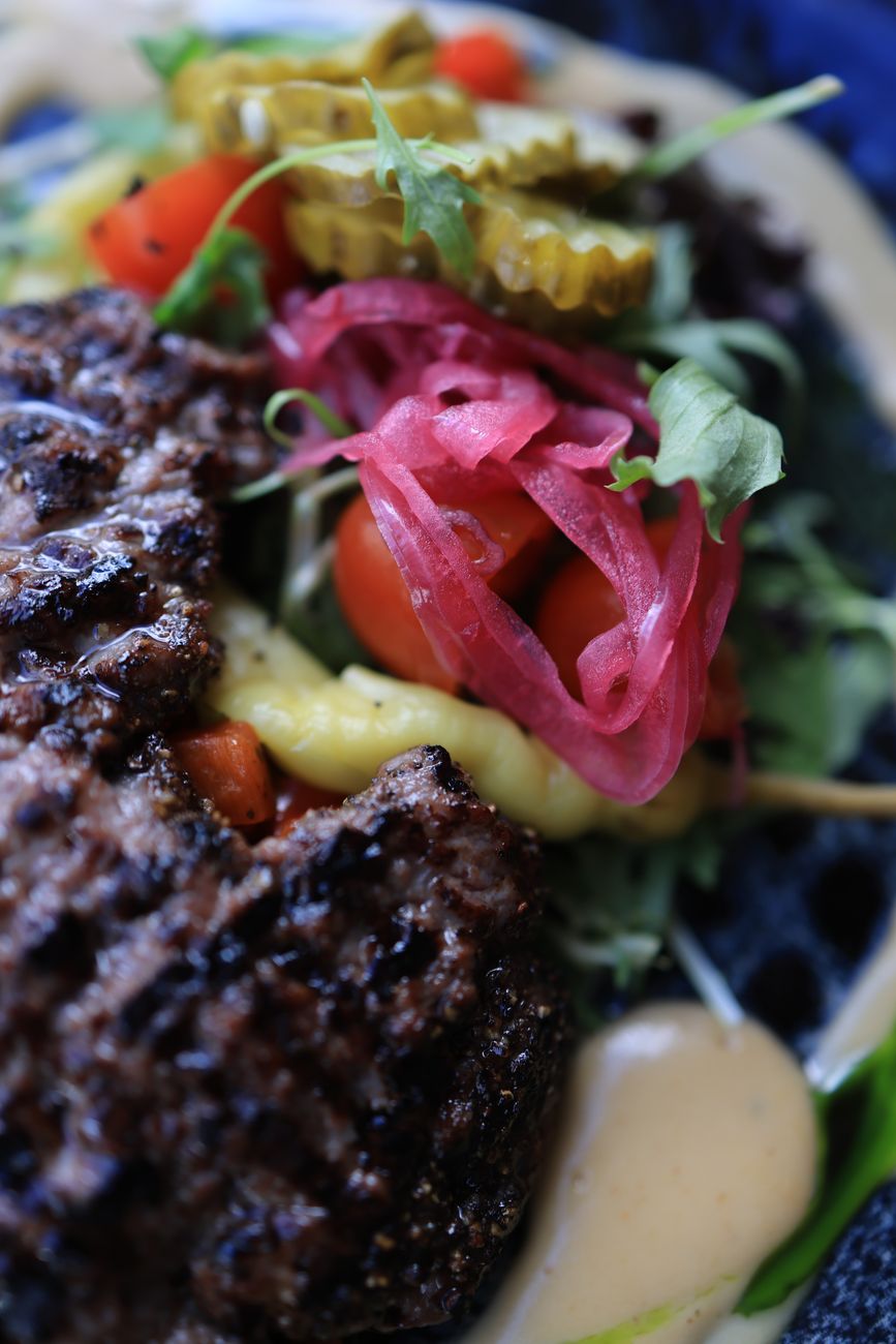 A beautifully assembled lamb shawarma burger in warm pita bread, topped with vibrant red harissa mayo, colorful pickled turnips, red onion slices, and fresh arugula, served on a rustic wooden plate