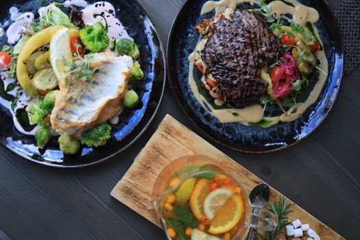 Top view of gourmet fish and steak dishes with fresh citrus and herbs on a stylish table setting.