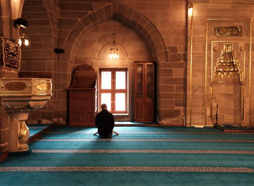 Free A man sitting and praying on a turquoise carpet inside a historic mosque, with ornate architecture and warm lighting. Stock Photo