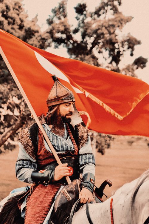 Free A medieval warrior in armor riding a horse, holding a red flag, in a historic outdoor setting. Stock Photo
