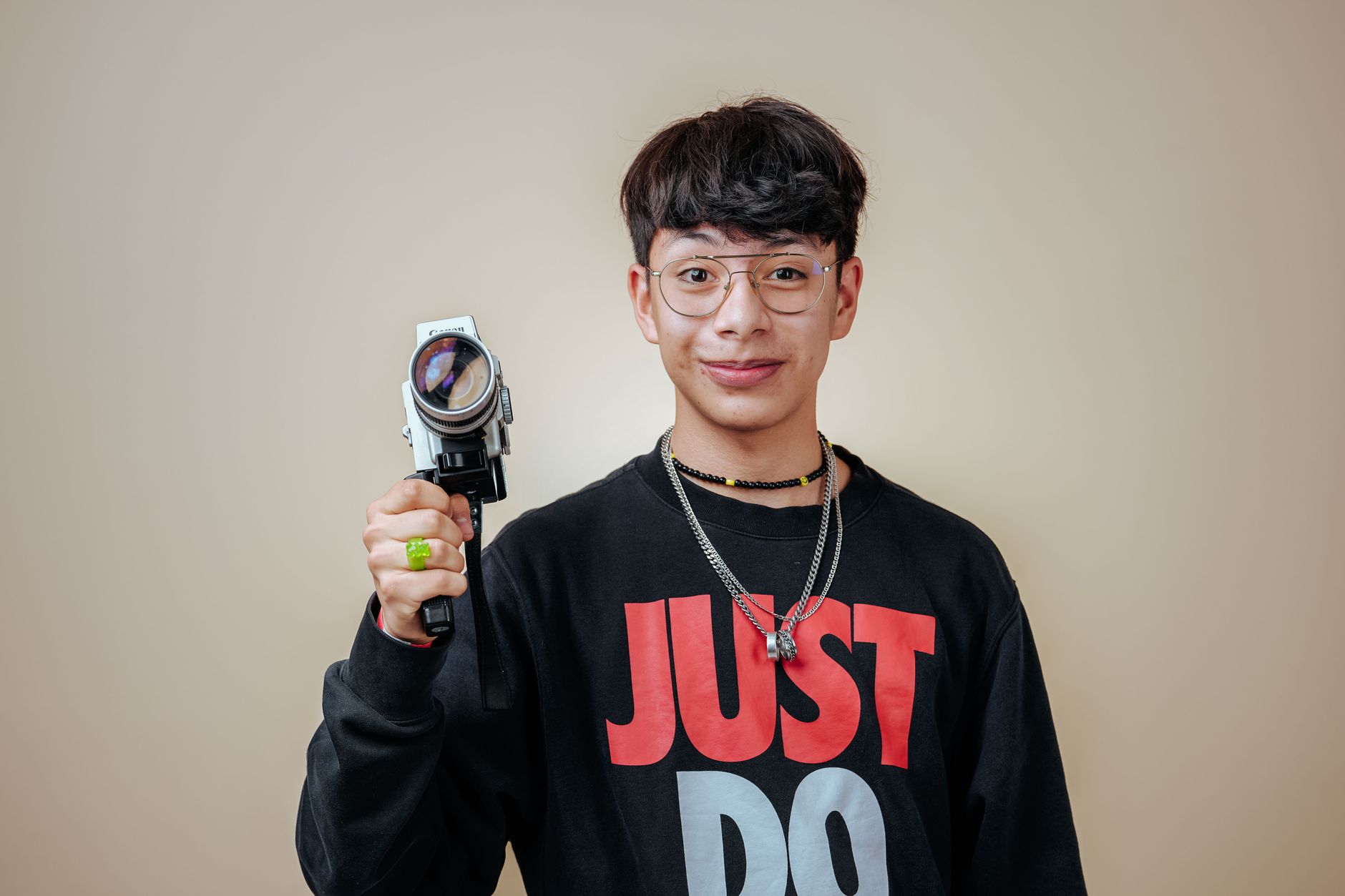A young man smiling while holding a vintage camera, wearing eyeglasses and a casual outfit.