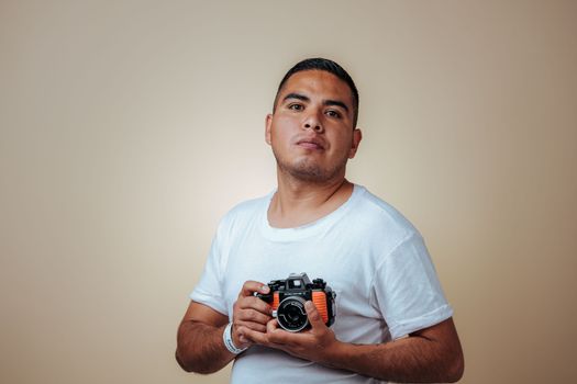 Portrait of a young man wearing a white t-shirt, holding a vintage camera against a beige studio background.